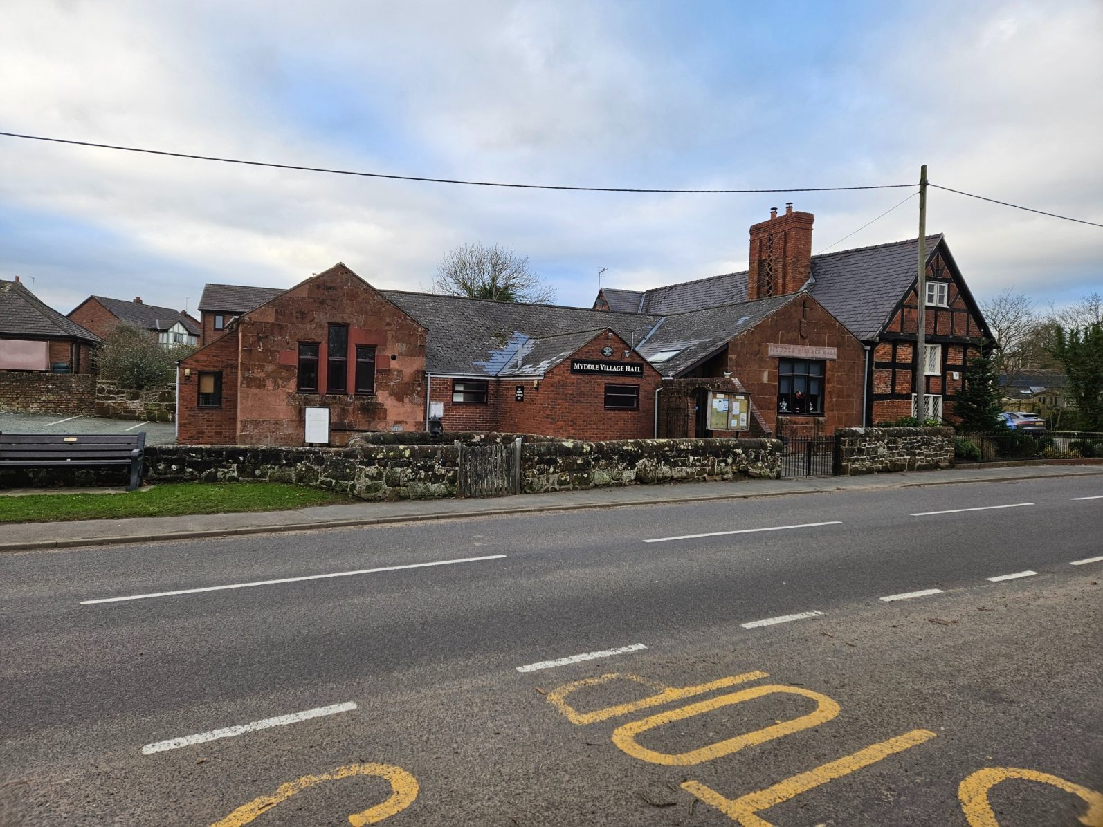 The hall from the road Historic brick building with a sloped roof, located by a road, partly surrounded by greenery.