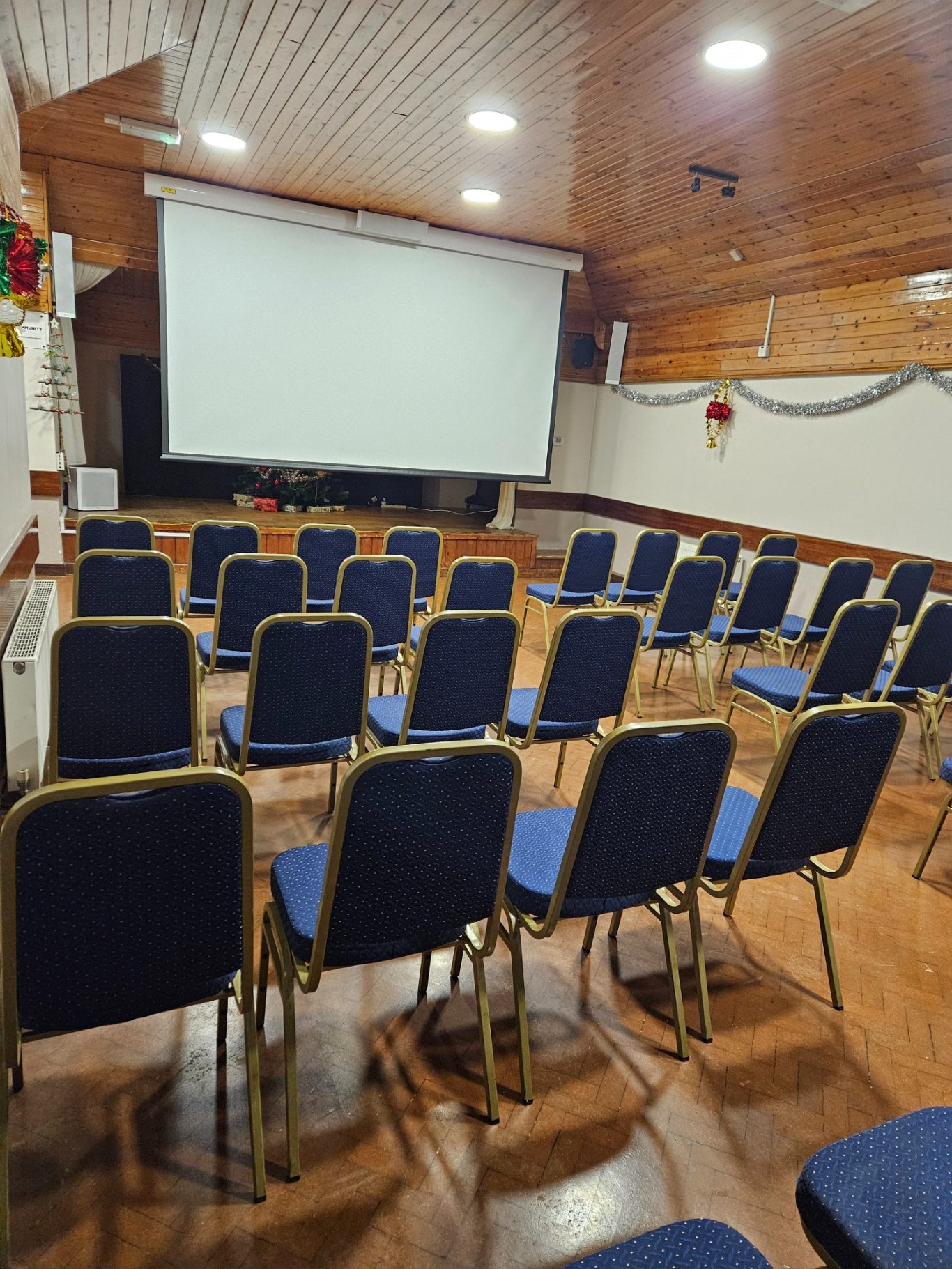 Screen and chairs set up for showing a film. Empty hall with rows of blue chairs facing a large projection screen.