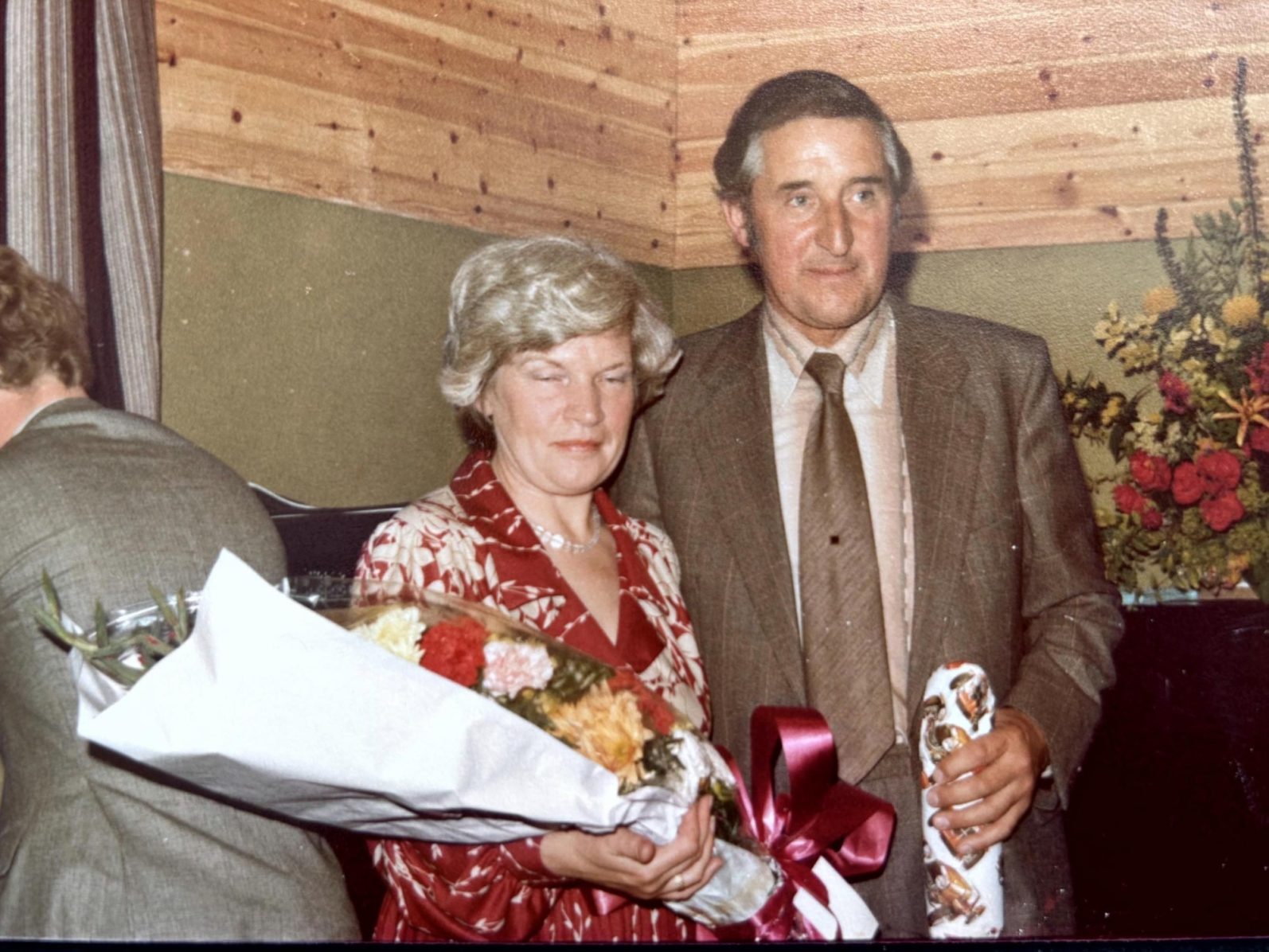 Mr and Mrs Boughey at the opening of the hall in 1980. A woman in a red floral dress holds a bouquet, standing with a man in a suit.